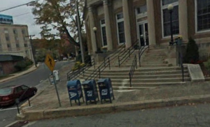 The Post Office facility at 91 Main St. has a steep staircase at its entrance with no ramp, lift, or other means for persons with limited mobility to get into the facility to conduct their business. For years, customers with disabilities could use a service ramp at the rear of the building; but the Postmaster recently suddenly shut down that option.  (Google Maps screenshot)